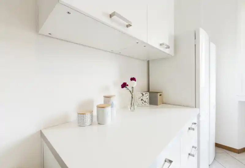 Bright minimalist kitchen counter with white cabinets, a refrigerator at the side and small decorative jars and a vase—clean and modern prep area.