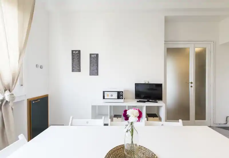 Bright, minimalist dining area with a white table, fresh flowers and a media console in the background; ideal for small gatherings and casual meals.