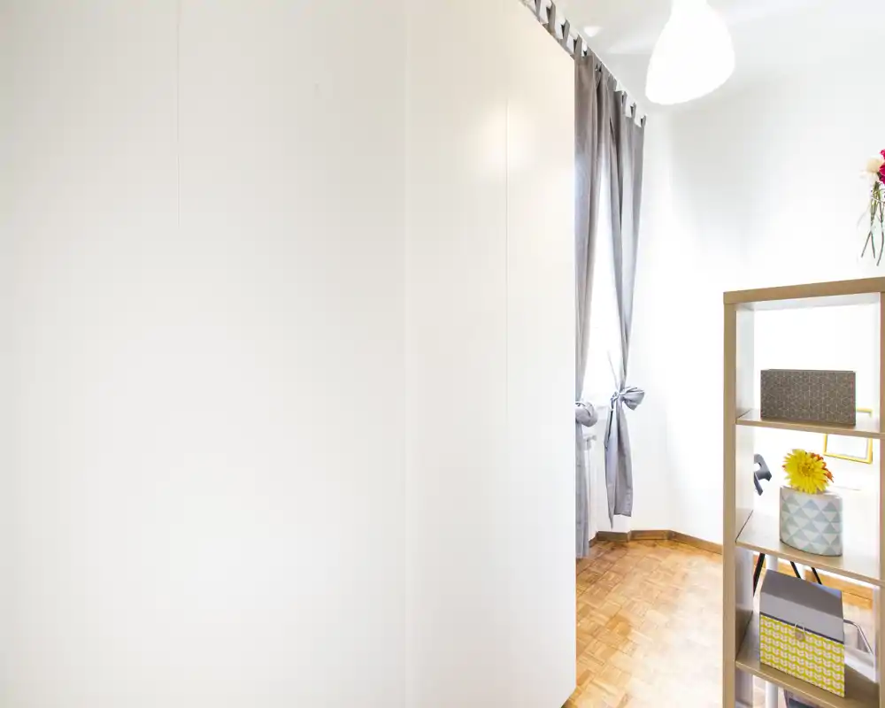 Bright, minimally furnished corner of a room with parquet flooring, a tall wardrobe or wall on the left, a window with tied curtains and an open shelving unit with decorative boxes and a vase.