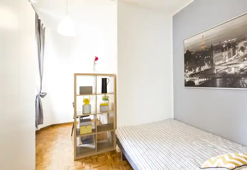 Bright, neatly staged bedroom with a striped single bed, small open shelving divider and framed wall art; natural light from a window creates an airy, welcoming feel.