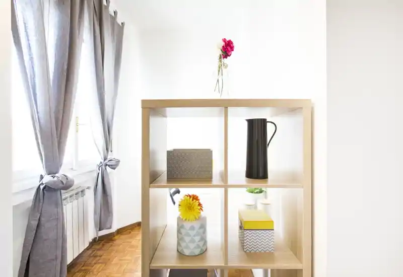 Bright, minimal living area corner with a wooden shelving unit, decorative vases and light-filled windows with grey curtains.
