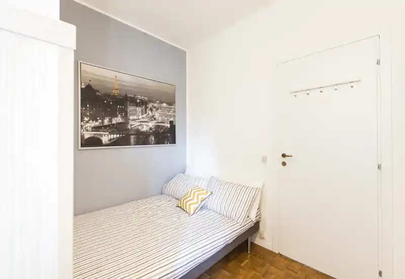 Small, bright bedroom with a double bed dressed in striped linens, a decorative yellow cushion and a framed cityscape above the headboard; white walls and parquet flooring create a clean, minimalist feel.