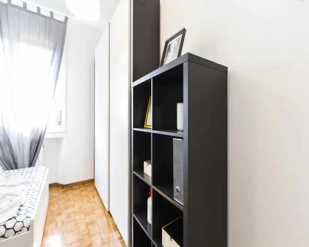 Angolo di camera luminoso e minimale con letto singolo vicino alla finestra, un armadio alto e uno scaffale a cubi nero per riporre oggetti.