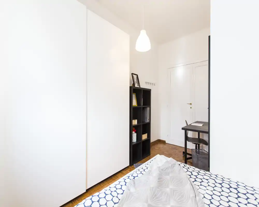 Camera da letto luminosa e moderna con copriletto a motivo, un grande armadio bianco e una scrivania angolare — stile pulito e minimal ideale per una persona.