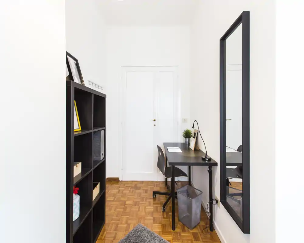 Piccolo studio/ingresso luminoso con scrivania nera minimal, sedia e scaffale. Pavimento in parquet, grande specchio a parete e dettagli decorativi semplici per un angolo lavoro funzionale.