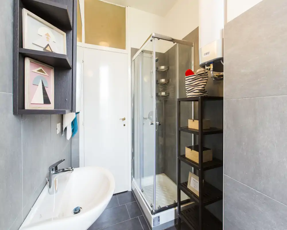 Modern compact bathroom with a glass shower enclosure, wall-mounted sink and open shelving; clean grey tiles and bright, even lighting.