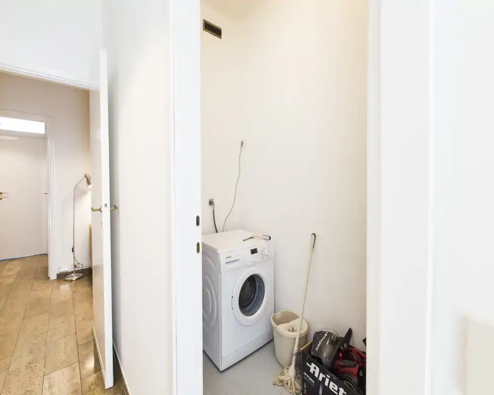 Small laundry/utility closet with a front-loading washing machine, mop, bucket and storage items in a white-painted alcove.