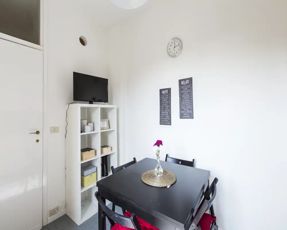 Compact dining area with a black table, four chairs and a decorative vase; bright white walls and shelving create a clean, modern look.