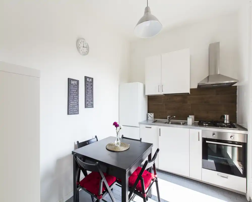 Bright, modern kitchenette with white cabinets, stainless appliances and a small dining table with red-cushioned chairs — ideal for a compact apartment.