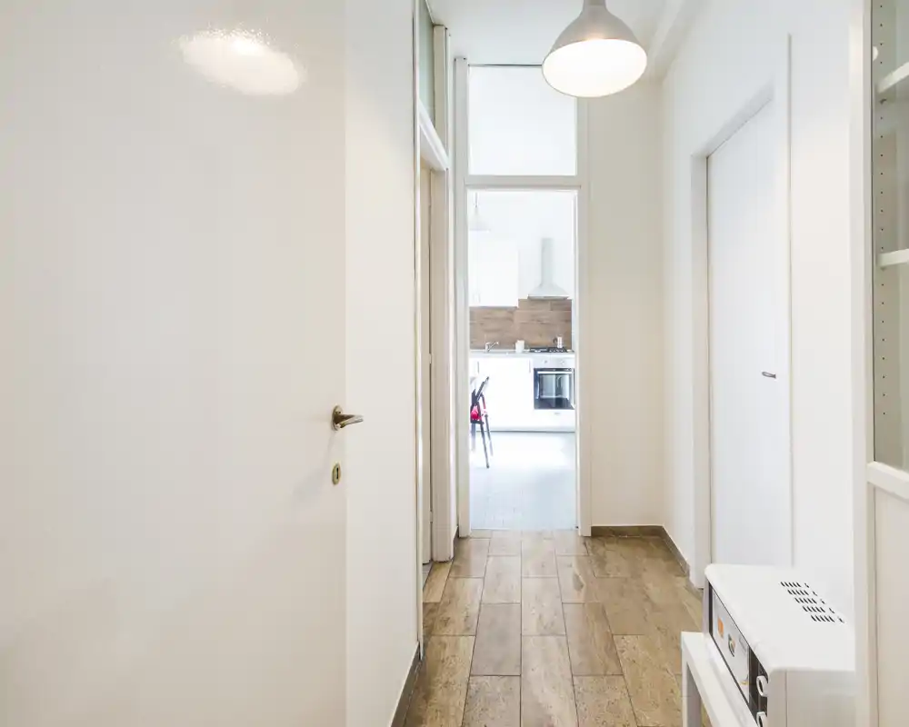 Bright, minimal hallway/entrance leading into a kitchen. White walls, wood-effect floor and pendant lighting create a clean, welcoming approach to the home.