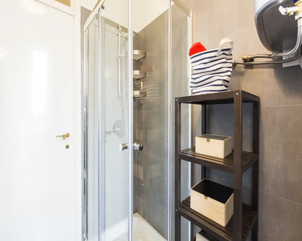 Compact modern bathroom with a glass corner shower, grey tiled walls and an open shelving unit for towels and storage.