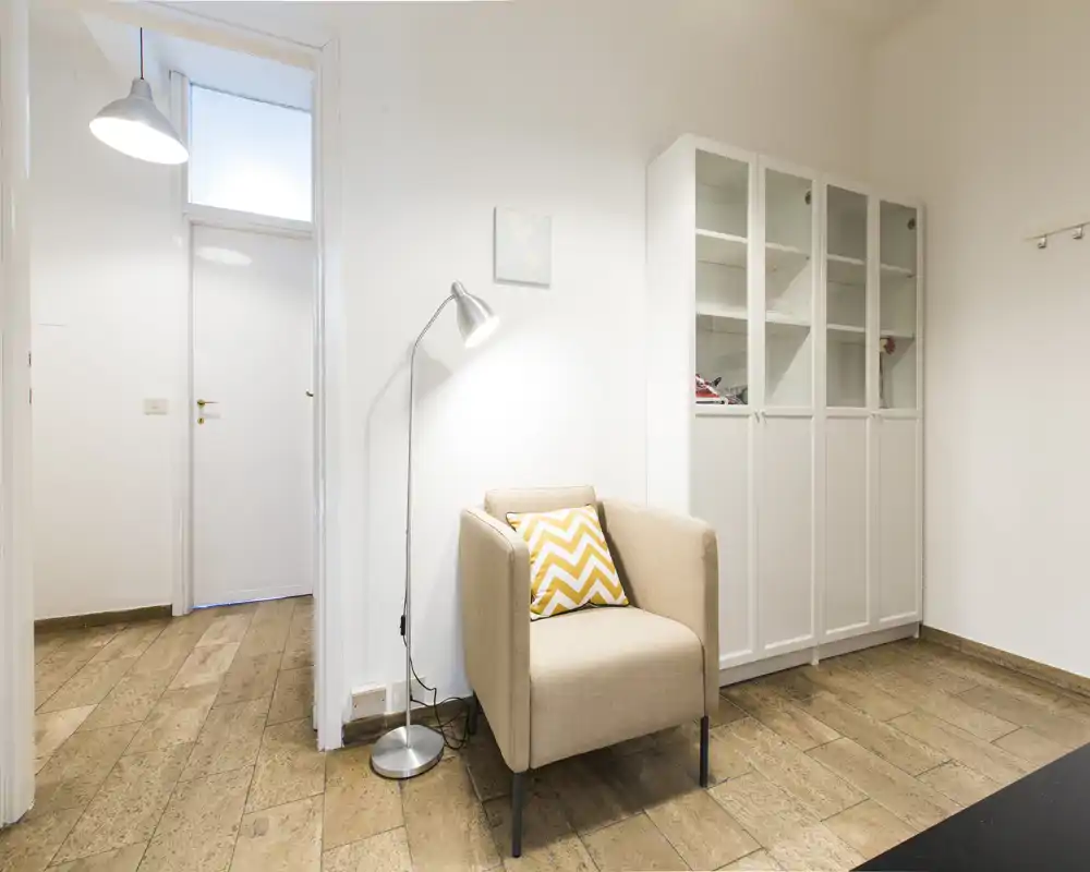 Compact, bright living corner with a comfortable armchair, patterned cushion and floor lamp beside a white display cabinet — ideal for relaxing or reading.