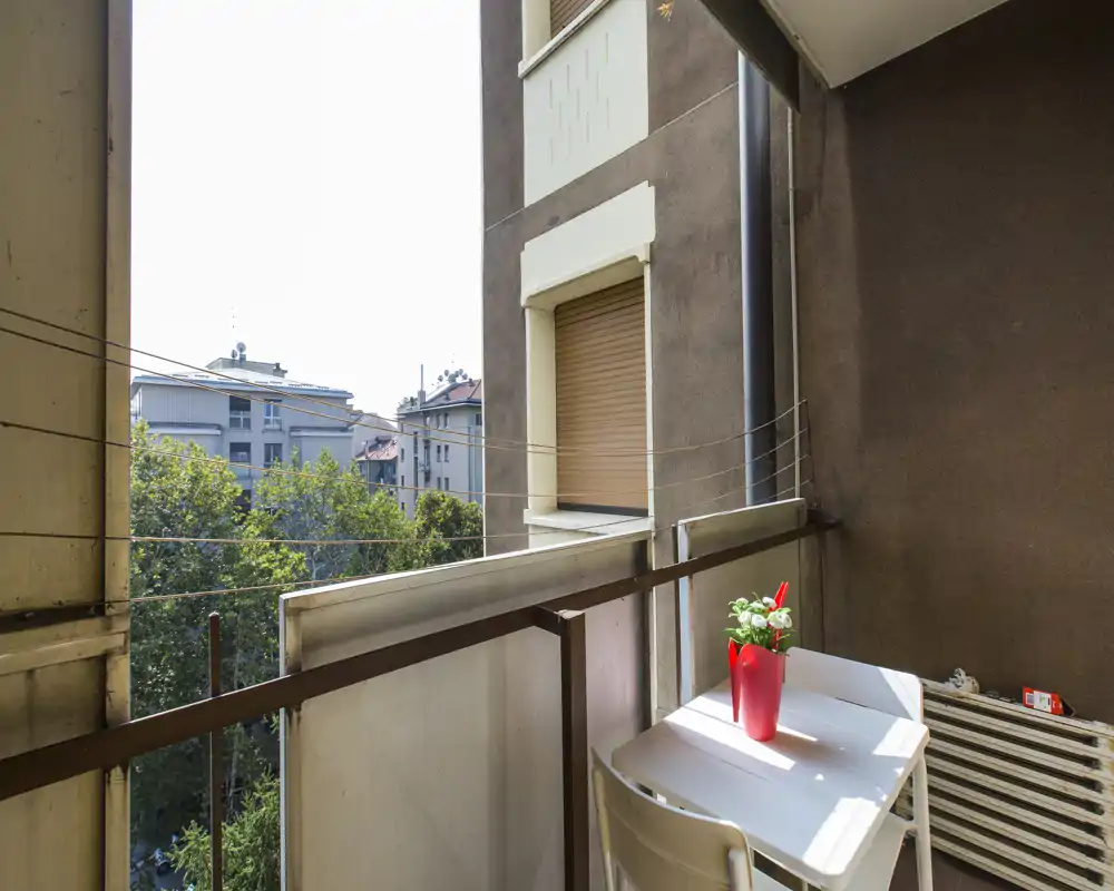 Sunny urban balcony with a small table and chair, perfect for a morning coffee. View includes nearby trees and apartment buildings.