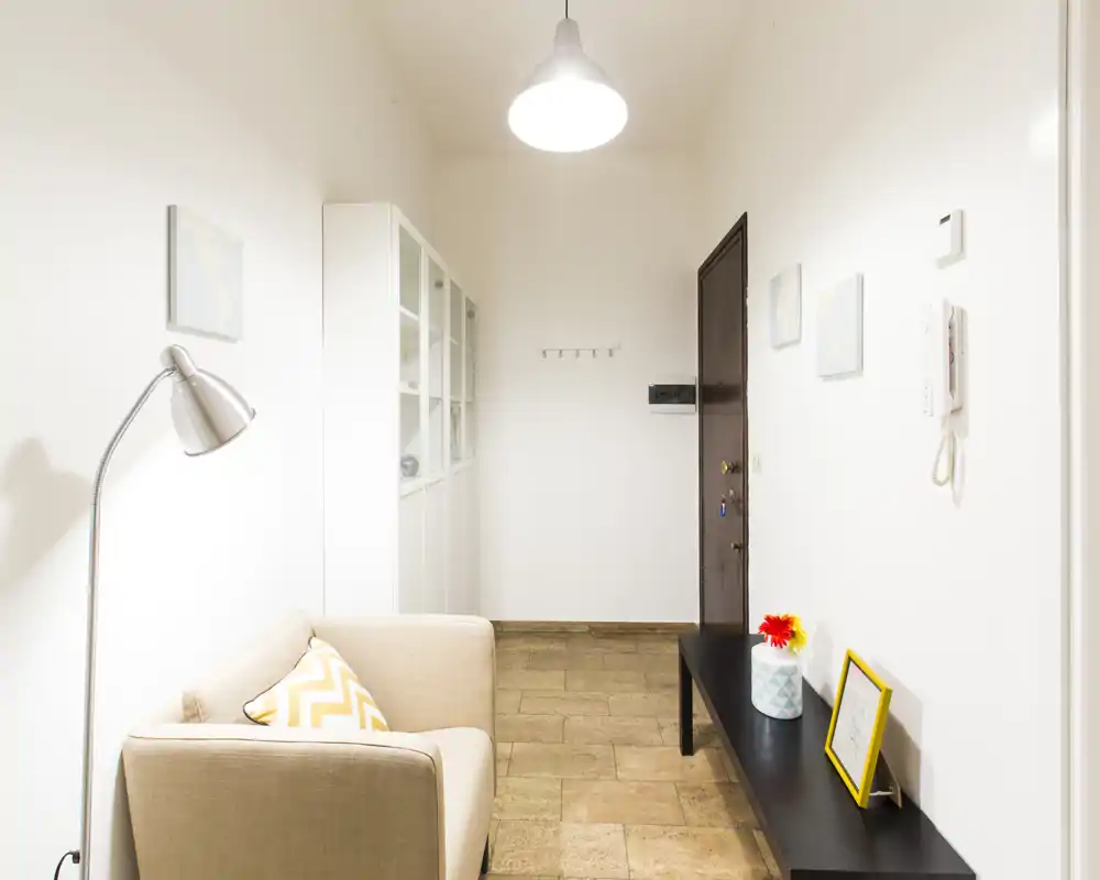 Bright, minimal entryway with a small sofa, floor lamp and a console table — clean white walls and tiled floor create a welcoming foyer.