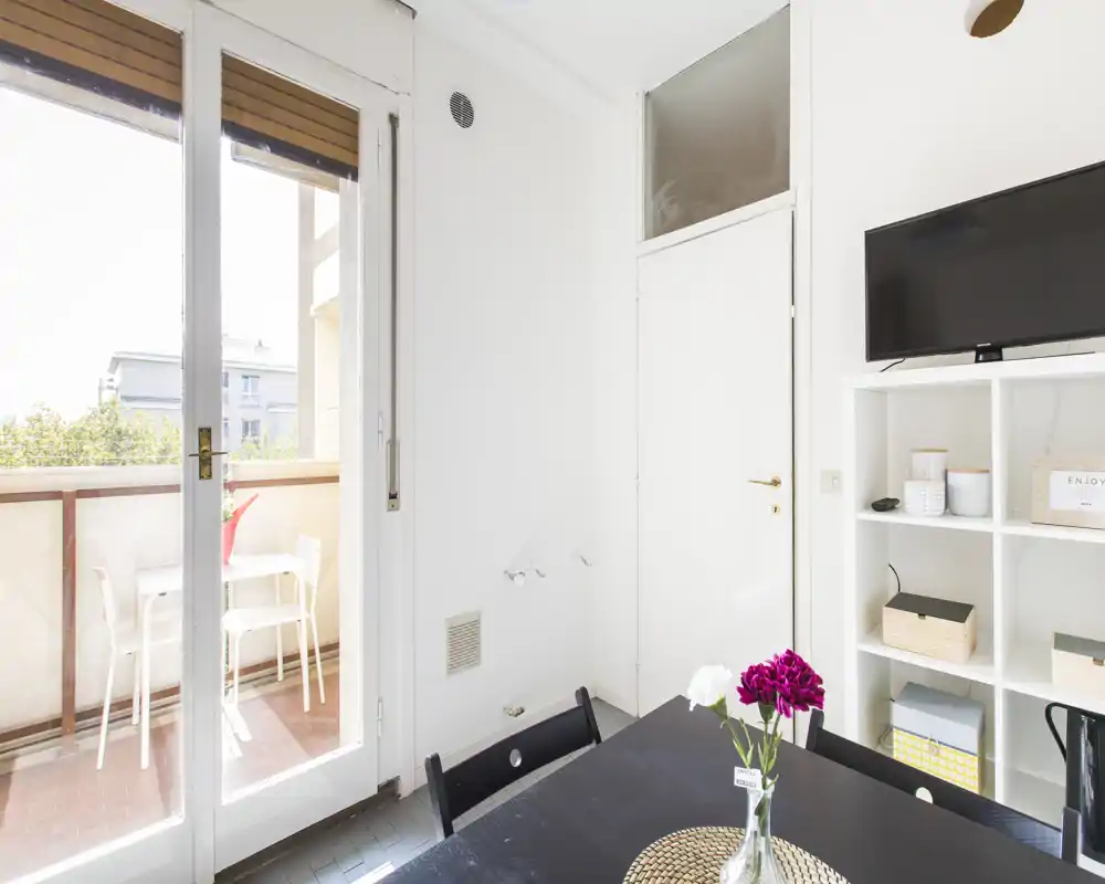 Bright, compact living area with a dining/work table, shelving and TV, opening onto a small balcony with outdoor seating.