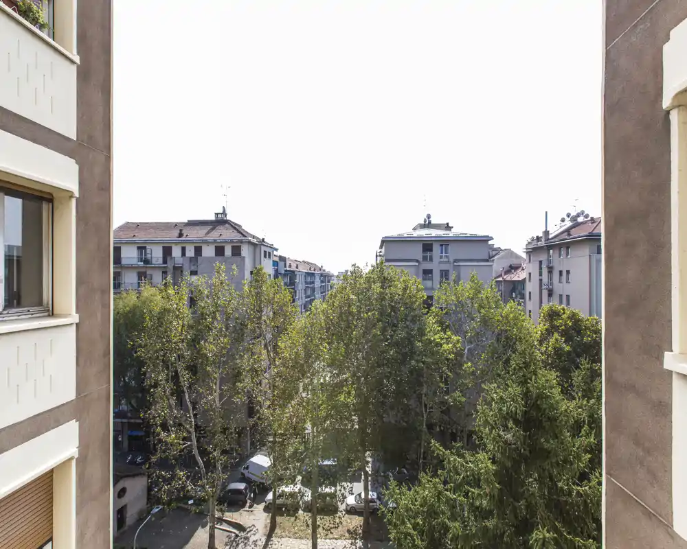 Sunny urban view from a window/balcony overlooking tree-lined courtyard and surrounding apartment buildings.