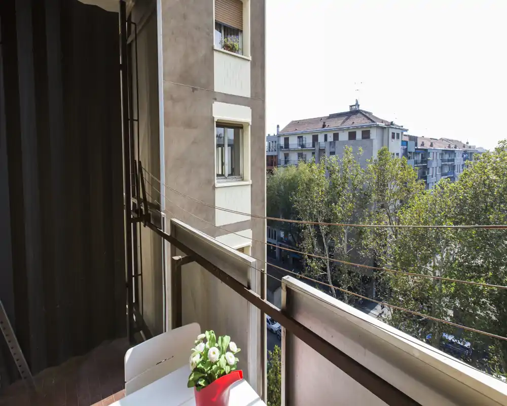 Sunny balcony with a small table and decorative plant, overlooking tree-lined street and neighboring buildings — ideal for morning coffee.