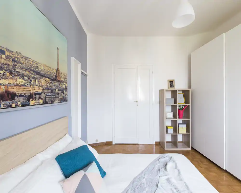 Dormitorio luminoso y moderno con una cama grande en primer plano, cojines decorativos y cabecero de madera; suelo de parquet y estantería sencilla para almacenamiento.