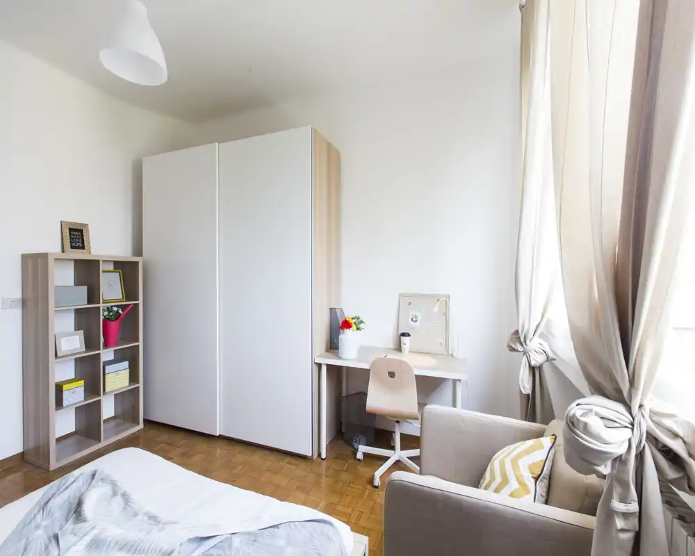 Dormitorio luminoso y minimalista con cama visible, armario, escritorio y sillón acogedor junto a una gran ventana — ideal para descansar o trabajar con luz natural.