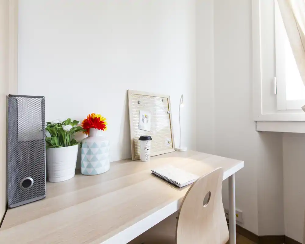 Espacio de trabajo luminoso y minimalista con escritorio de madera clara, una pequeña planta y jarrón decorativo, panel de corcho y libreta — ideal para estudiar o trabajar desde casa.
