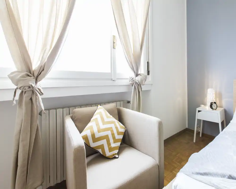Rincón de dormitorio luminoso y acogedor con un sillón, cojín decorativo de chevron y mesita de noche con lámpara. La gran ventana aporta abundante luz natural y tonos neutros que crean calma.
