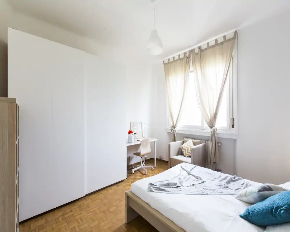 Dormitorio luminoso y minimalista con cama doble, gran armario, escritorio y un cómodo sillón junto a la ventana que aporta mucha luz natural.