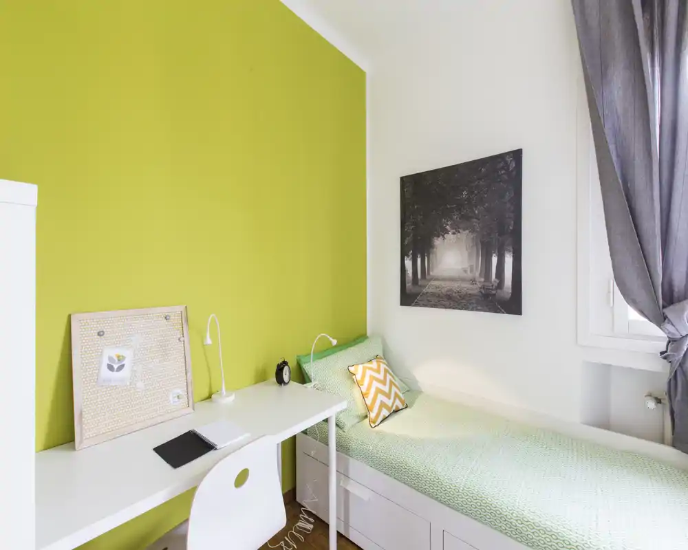 Acogedora habitación individual con escritorio blanco, cama con detalles verdes y mucha luz natural desde la ventana — ideal para estudiante o persona sola.