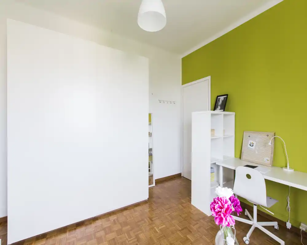 Rincón de habitación luminoso y minimalista con escritorio blanco, estantería, silla con ruedas y suelo de parquet junto a una pared verde; no se aprecia una cama ni zona de descanso clara.