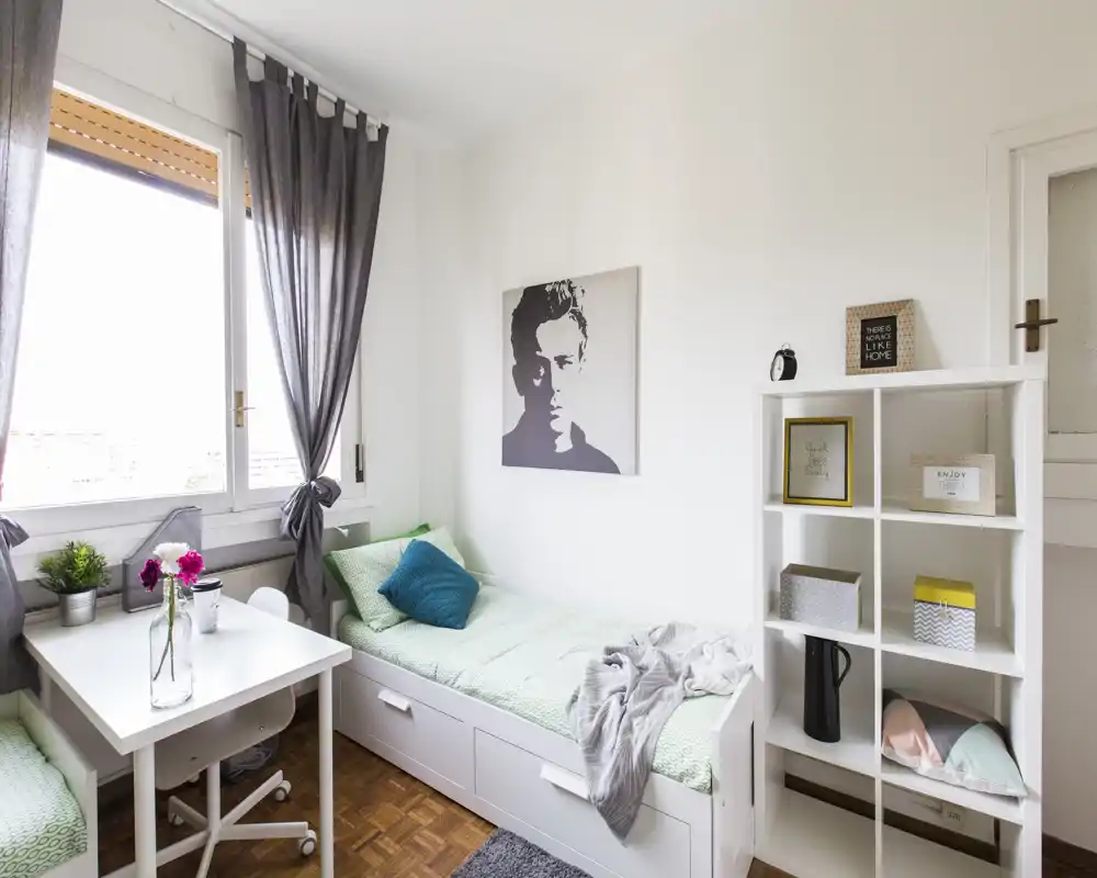 Dormitorio pequeño y luminoso con cama individual, ropa de cama verde claro y cojines decorativos. Incluye un escritorio junto a la ventana, estanterías para almacenamiento y una decoración moderna y minimalista ideal para una persona.