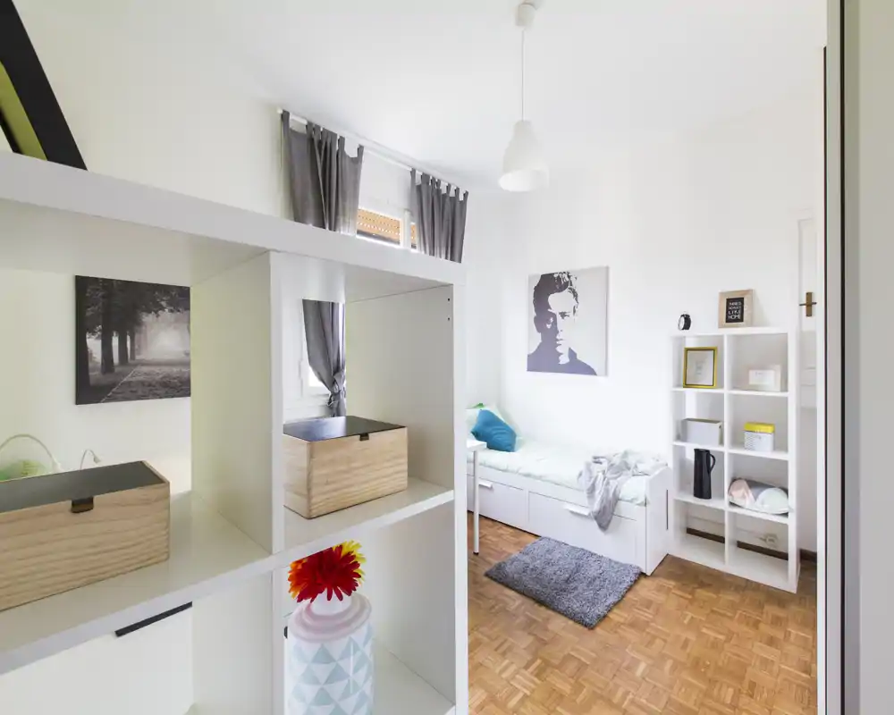 Habitación individual luminosa y bien presentada con una cama blanca, textiles suaves y una pequeña estantería; luz natural y decoración minimalista que generan un ambiente acogedor.