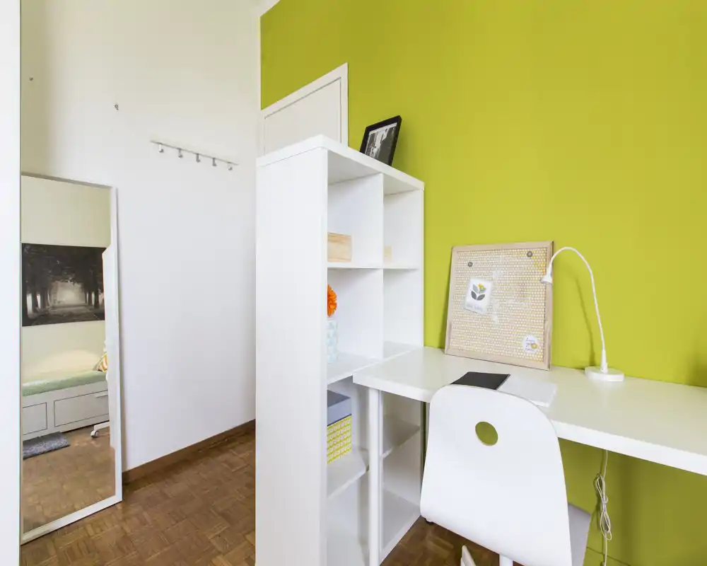 Rincón de dormitorio compacto con escritorio blanco y estantería frente a una pared verde brillante; un espejo de cuerpo entero a la izquierda refleja la cama en la habitación contigua. El espacio de trabajo es luminoso y ordenado, ideal para estudiar o trabajar desde casa.