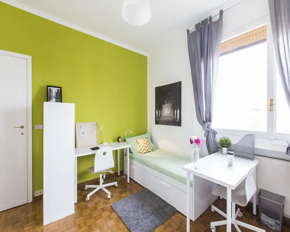 Dormitorio individual luminoso y bien decorado con cama blanca, escritorio y una ventana grande que aporta mucha luz natural — ideal para estudiante o persona sola.