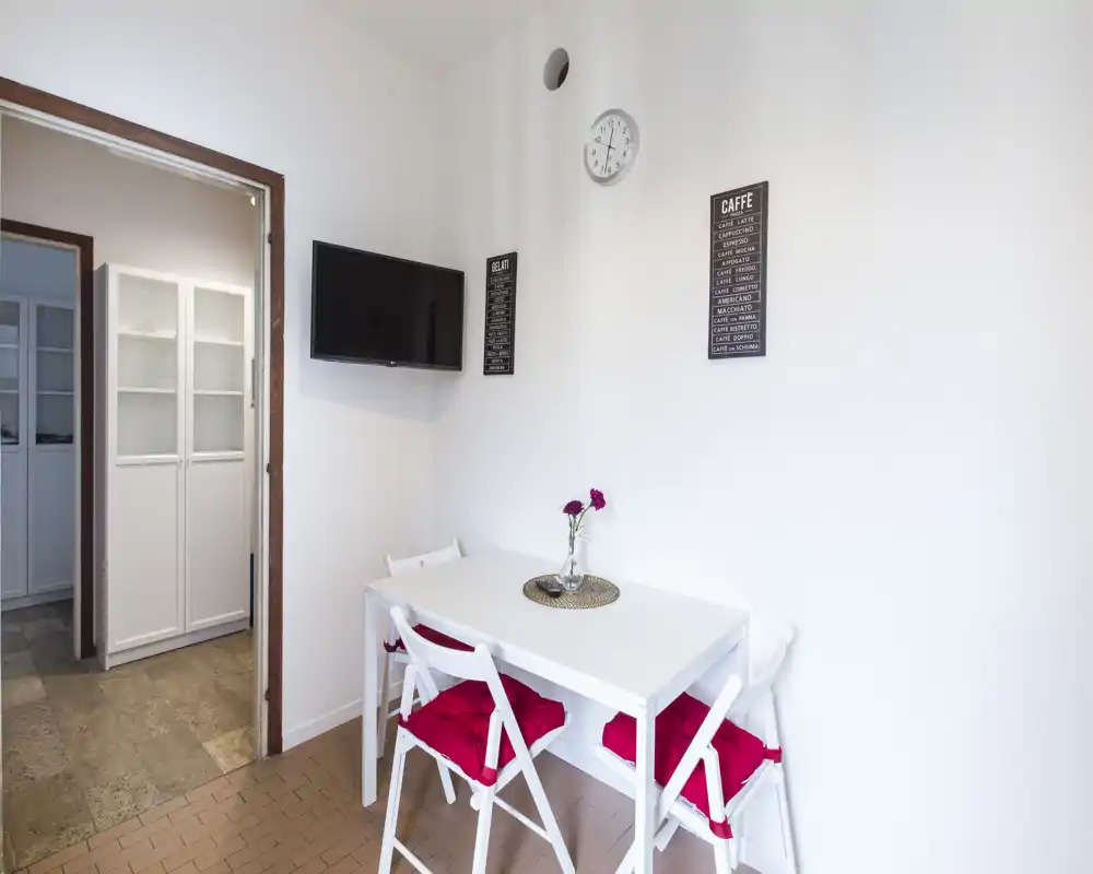 Rincón comedor compacto con mesa blanca y cuatro sillas plegables con cojines rojos, un pequeño jarrón con flores y una TV en la pared — luminoso, sencillo y bien organizado para comidas informales.