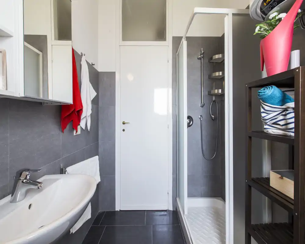 Baño moderno y limpio con lavabo, armario con espejo, ducha cerrada y estantería con toallas. Baldosas grises y puerta blanca crean una apariencia fresca y contemporánea ideal para alquileres.