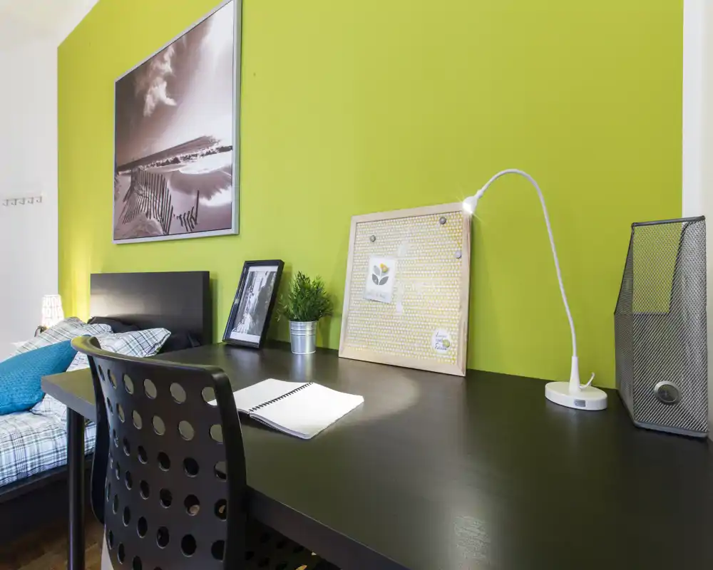 Rincón de dormitorio moderno y luminoso con escritorio: área de trabajo ordenada frente a la cama, pared verde llamativa y decoración contemporánea.