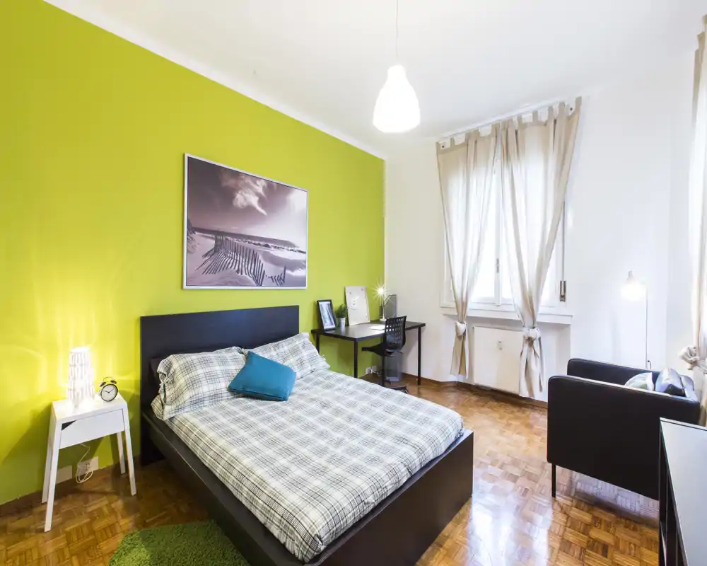 Dormitorio moderno y luminoso con cama doble, mesita, escritorio y zona de estar; pared verde lima y gran ventana que crean un espacio acogedor y ventilado.