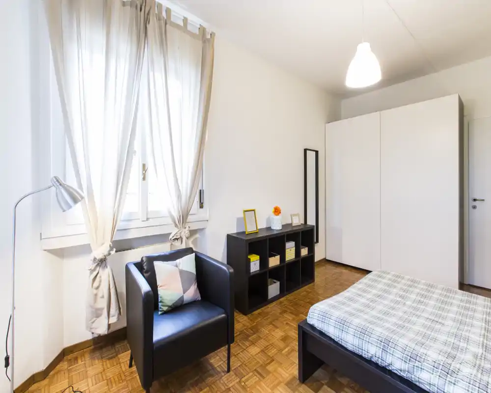 Dormitorio moderno y luminoso con cama doble, armario y un sillón acogedor junto a la ventana; suelo de parquet y suave luz natural.