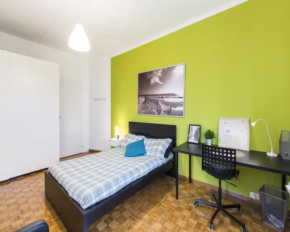 Dormitorio moderno y luminoso con cama doble, ropa de cama a cuadros y cojín turquesa. Un largo escritorio con silla y lámpara frente a una pared verde brillante, armario empotrado y suelo de parquet.
