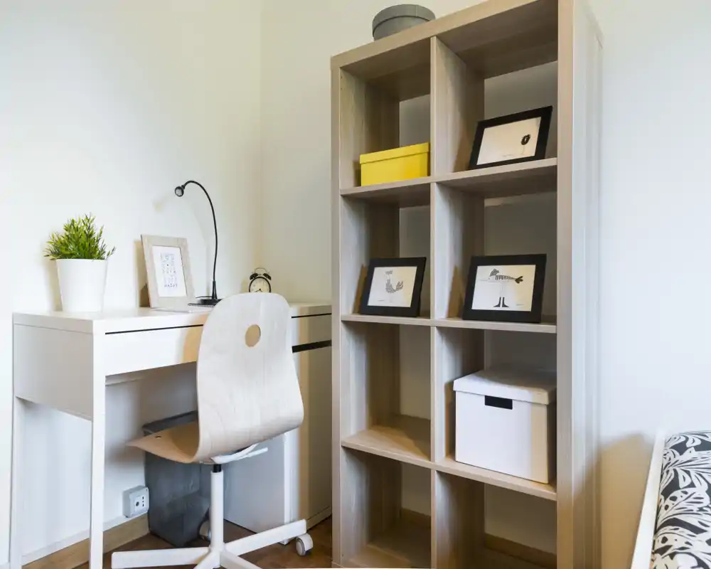 Bright, minimal bedroom corner with a white desk, modern wooden chair and open shelving. The composition highlights a compact workspace and storage beside a partially visible bed, ideal for listings emphasizing a study nook.