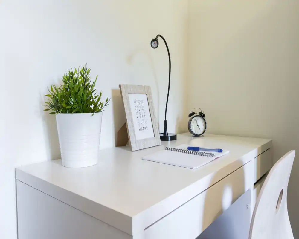 Bright, minimalist desk corner with a white table, decorative plant, framed print, desk lamp and an alarm clock — a tidy workspace suitable for remote work.