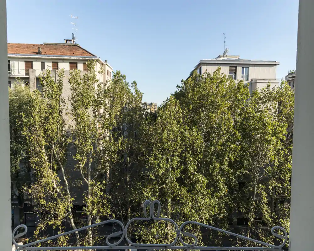 Vista dal balcone con alberi rigogliosi e palazzi circostanti sotto un cielo sereno — un tranquillo panorama urbano.