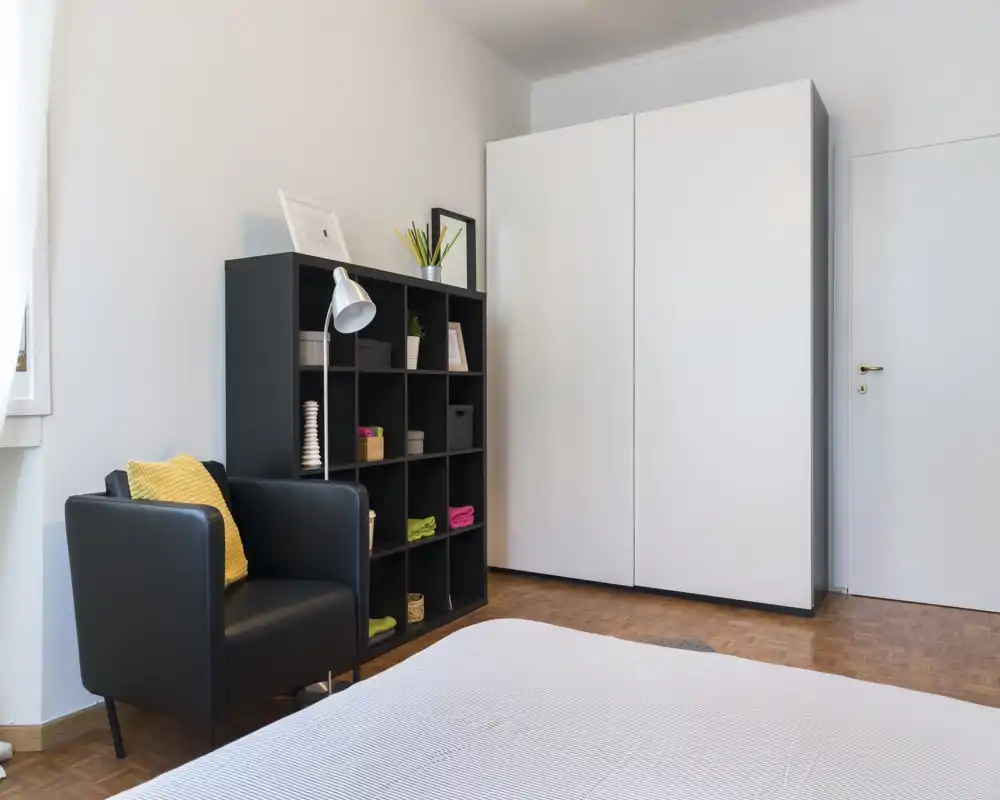 Camera da letto moderna e luminosa con letto ben visibile in primo piano, poltrona nera con cuscino giallo, scaffalatura aperta e grande armadio bianco — spazio ordinato e ideale per un annuncio immobiliare.