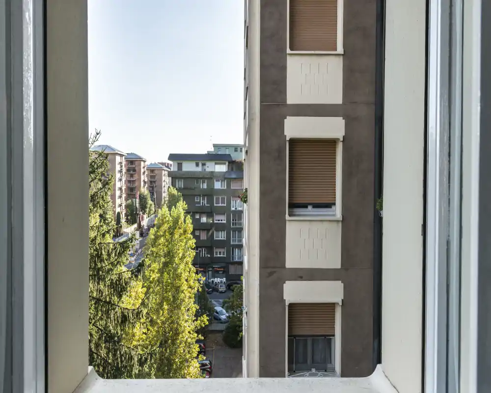 Vista su strada e cortile inquadrata da una finestra, con palazzi vicini e alberi folti sotto una luce diurna luminosa.