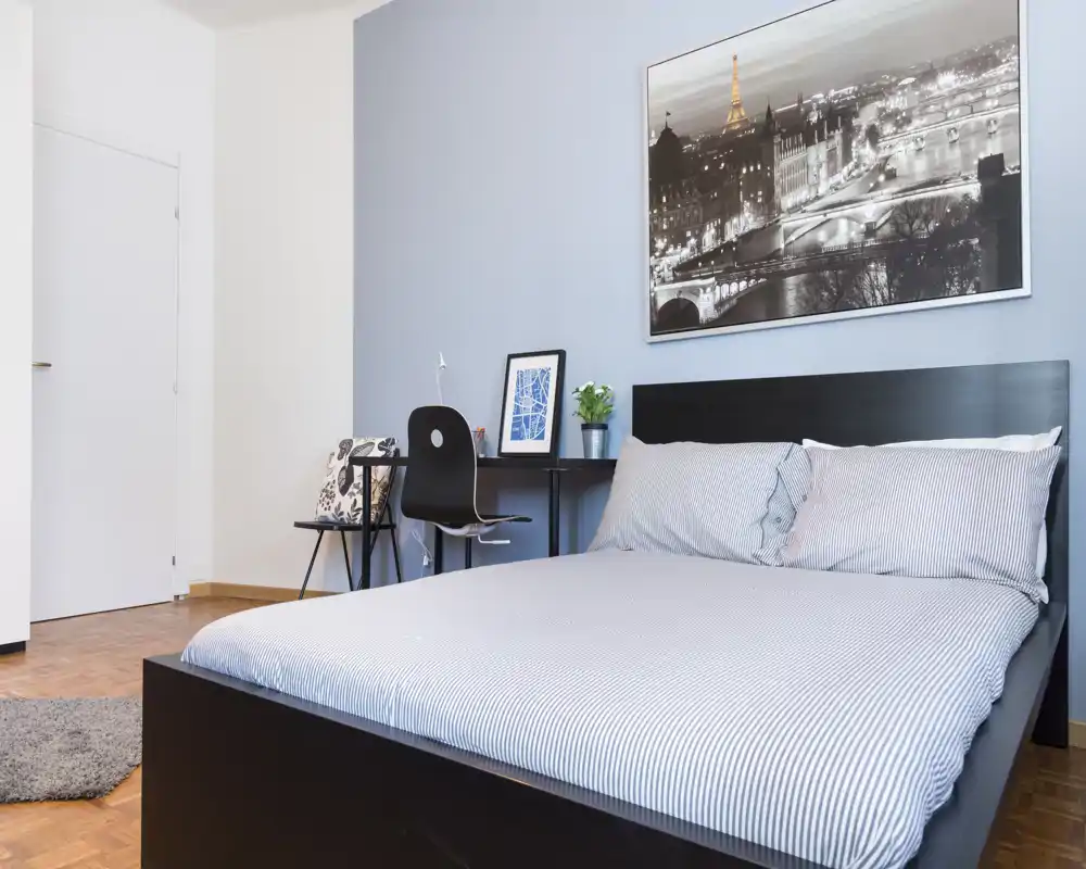 Camera da letto luminosa e moderna con un letto matrimoniale rifatto, una scrivania con sedia e un elegante quadro in bianco e nero sopra la testiera.