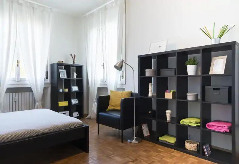 Camera da letto moderna e luminosa con letto basso, scaffali modulari e una poltrona comoda vicino alla finestra. Finestre grandi con tende leggere assicurano luce naturale e soluzioni di stoccaggio ordinate mantengono lo spazio pulito.