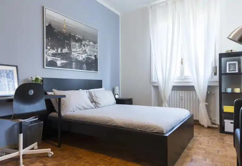 Camera da letto moderna e luminosa con letto matrimoniale, scrivania e scaffale; luce naturale morbida dalla finestra e arredi minimal rendono l'ambiente accogliente.