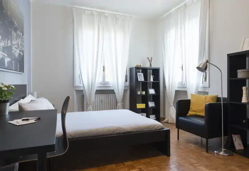 Camera da letto luminosa e ben arredata con letto matrimoniale al centro, luce naturale da due finestre alte, una scrivania, scaffalature e una poltrona accogliente con cuscino decorativo.