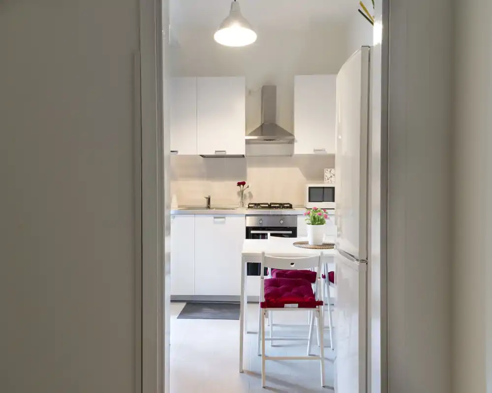 Cucina moderna e luminosa con mobili bianchi, piano cottura a gas, forno e un piccolo tavolo con cuscini rossi — ambiente ordinato e ben illuminato, ideale per appartamenti compatti.