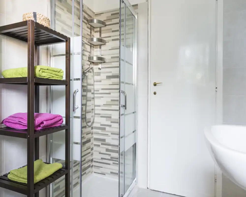 Bagno moderno e pulito con box doccia in vetro, piastrelle decorative e scaffale aperto con asciugamani colorati.