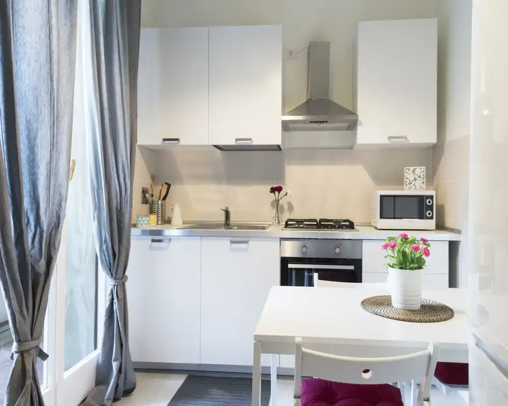 Angolo cottura moderno e luminoso con mobili bianchi, piano cottura a gas e un piccolo tavolo da pranzo. La luce naturale dalla porta e gli elementi decorativi (fiori e cuscini) lo rendono accogliente.